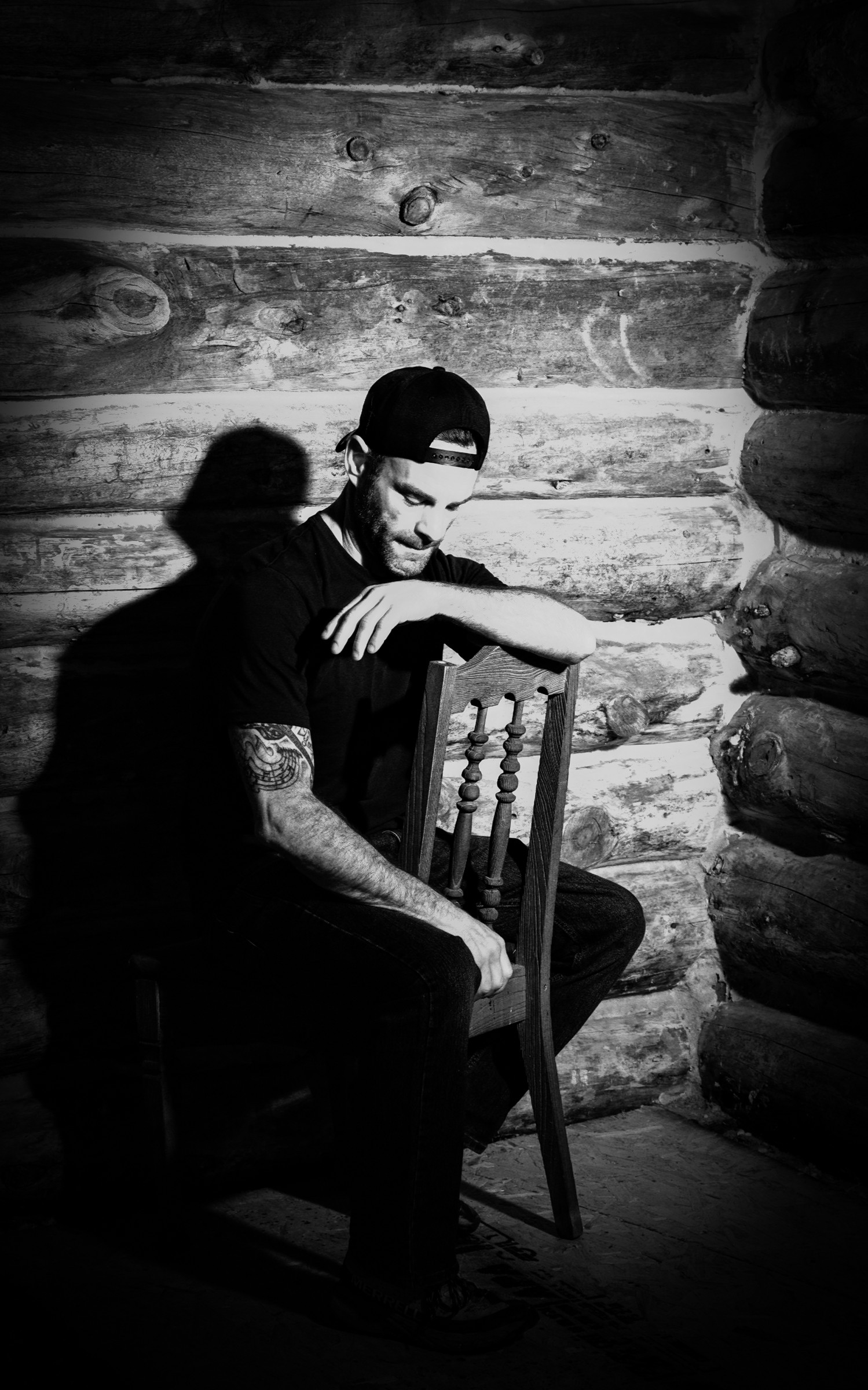 Black and white portrait of a seated man beside a rustic wooden wall