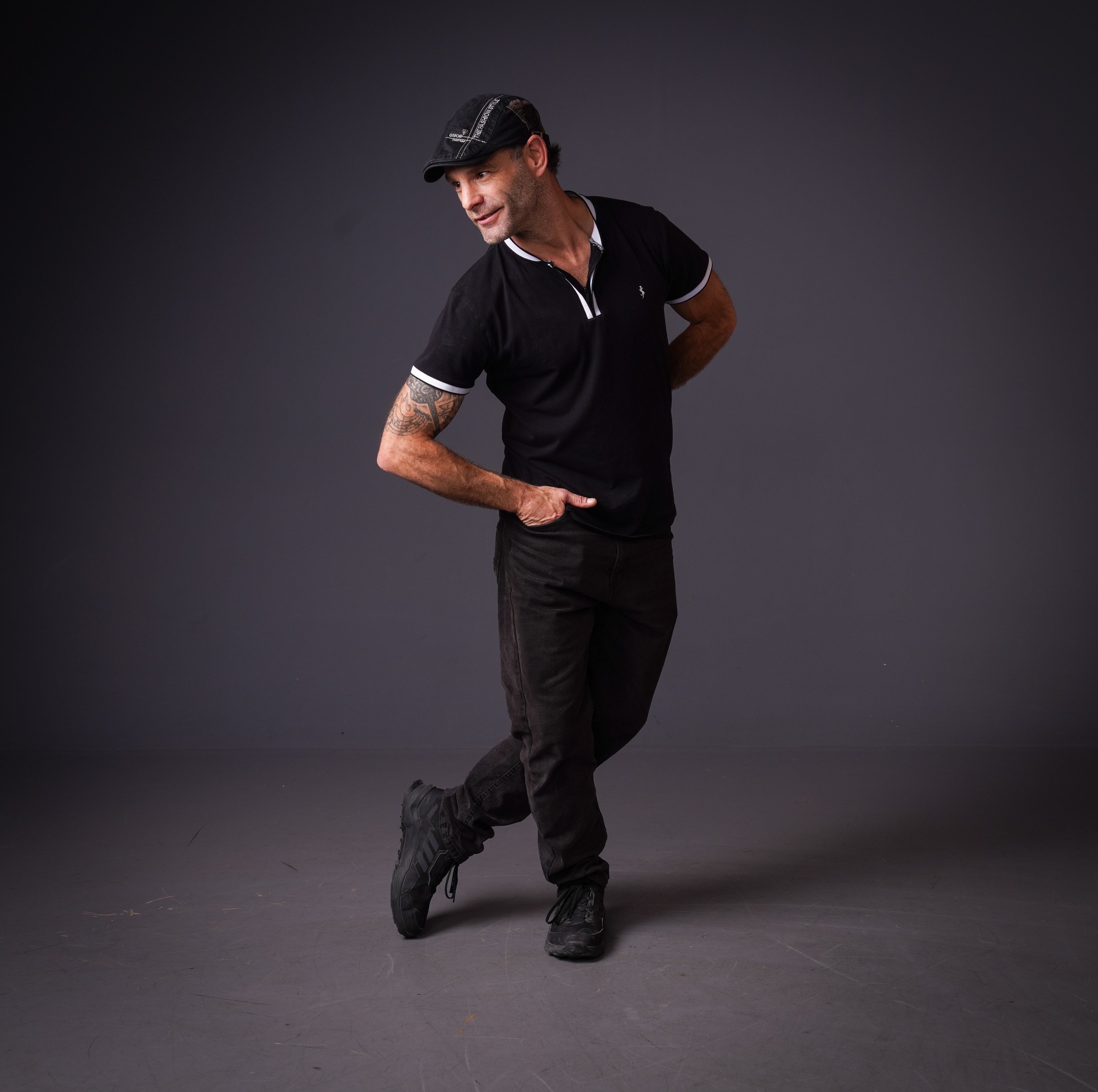 Man in black clothing and a cap striking an energetic pose against a dark backdrop